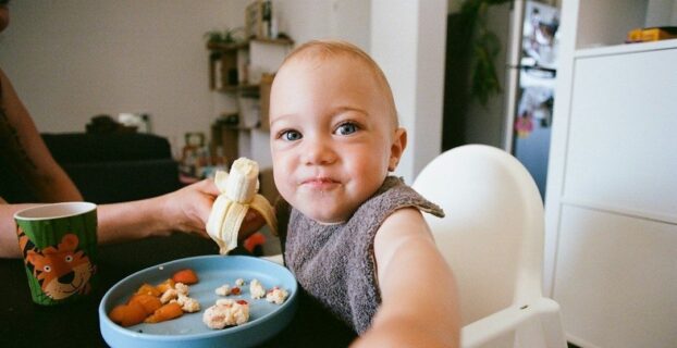 Småbarn smiler i barnestol med banan og mat på tallerkenen